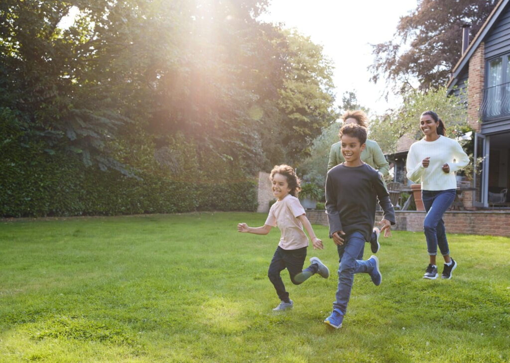 A mother, father, and two children running on a grassy lawn in a backyard, with sunlight streaming through the trees, and a house in the background.