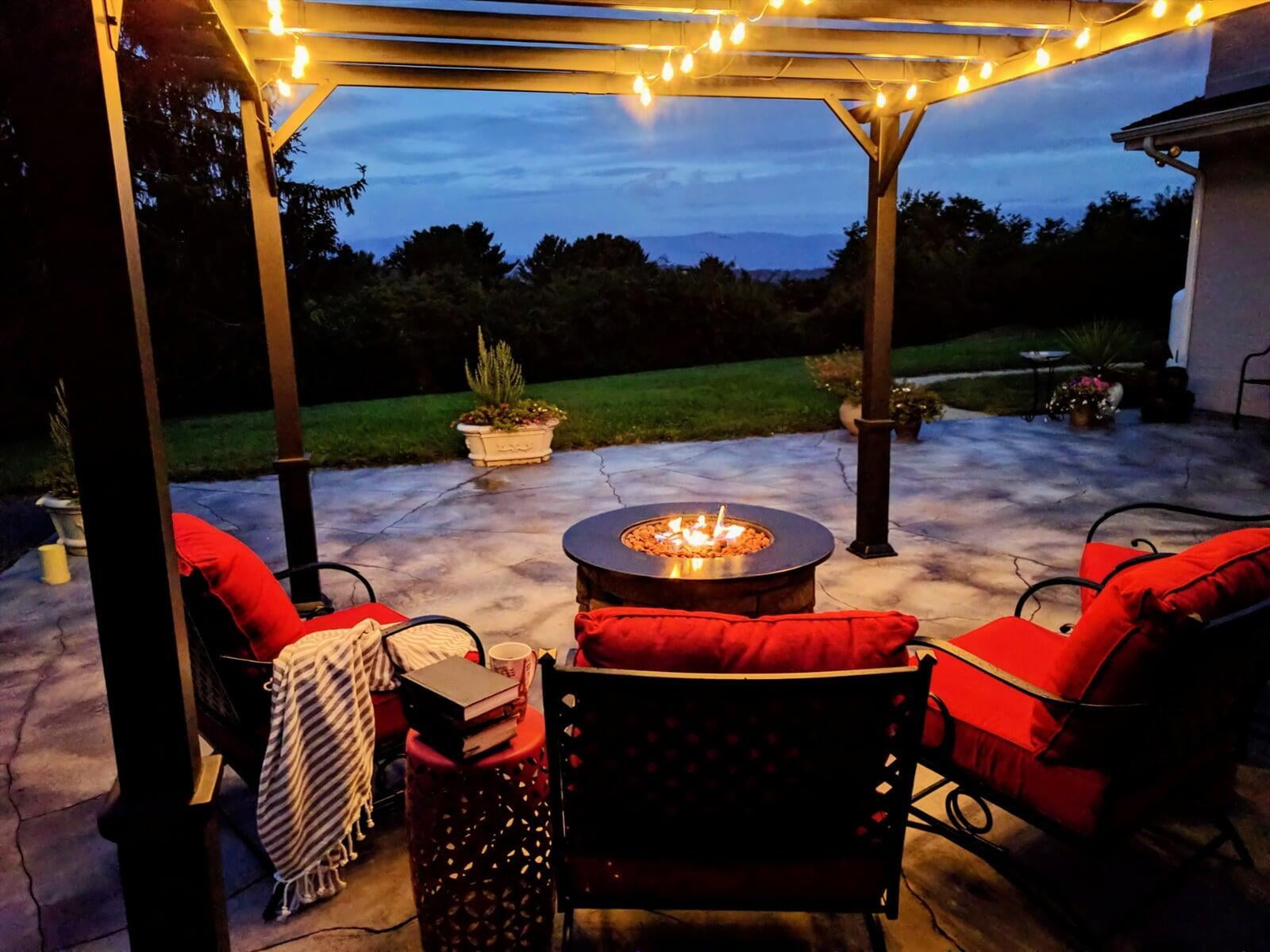 Outdoor living space with a gas fire pit, red-cushioned chairs, and string lights hanging from a pergola, photographed at dusk.